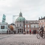 Aged masonry cathedral and sculpture near buildings on tiled pavement with walking people in Copenhagen